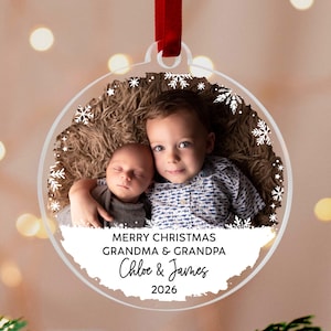 Pode incluir: Um enfeite de Natal circular em acrílico transparente com uma fotografia de duas crianças, um bebé e uma criança pequena, rodeado por uma borda de flocos de neve brancos. O texto diz MERRY CHRISTMAS GRANDMA & GRANDPA, Chloe & James, 2026.