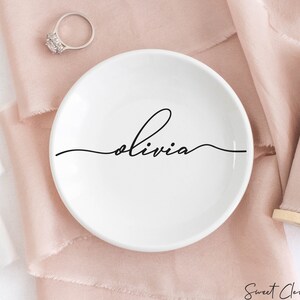 May include: White ceramic ring dish with the name "Olivia" written in cursive on the front. The dish is sitting on a pink fabric surface.
