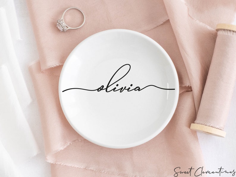 May include: White ceramic ring dish with the name "Olivia" written in cursive on the front. The dish is sitting on a pink fabric surface.