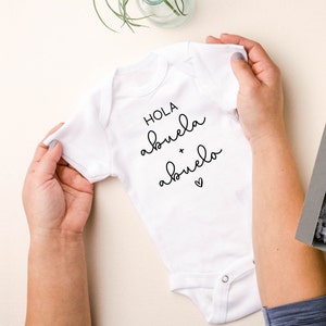 Op de afbeelding: Witte baby romper met zwarte tekst die "Hola abuela + abuelo" met een klein hartje zegt.
