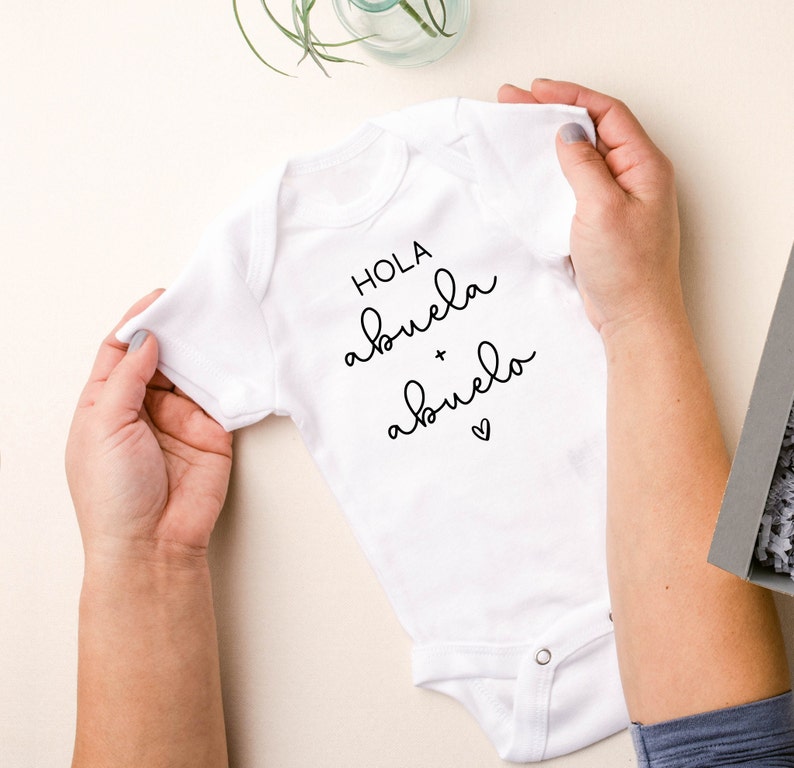 Op de afbeelding: Witte baby romper met zwarte tekst die "Hola abuela + abuelo" met een klein hartje zegt.