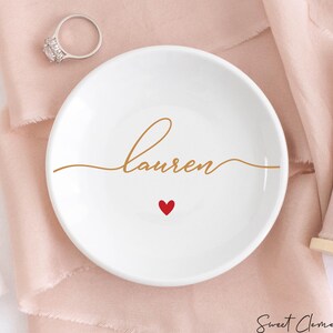 K&ouml;nnte beinhalten: Eine wei&szlig;e Keramik-Ring Schale mit dem Namen "lauren" in goldener Schrift und einem kleinen roten Herzen.