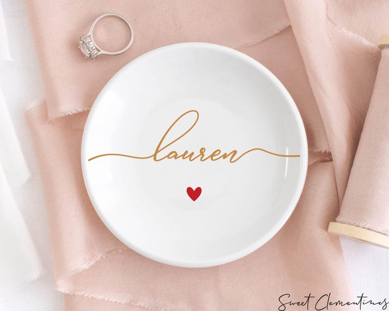 K&ouml;nnte beinhalten: Eine wei&szlig;e Keramik-Ring Schale mit dem Namen "lauren" in goldener Schrift und einem kleinen roten Herzen.