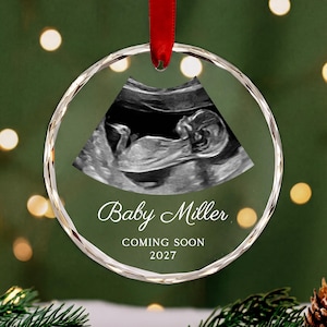 May include: A circular clear glass ornament with a faceted edge, featuring a black and white ultrasound image of a fetus. The text "Baby Miller" is printed in white script, followed by "COMING SOON" and "2027" in a clean white font.