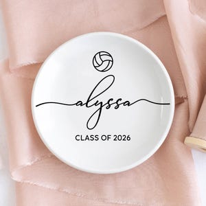 May include: A white ceramic ring dish with a black volleyball graphic and the name "alyssa" in cursive script. Below the name is the text "CLASS OF 2026". The dish is round and sits on a pink fabric surface.