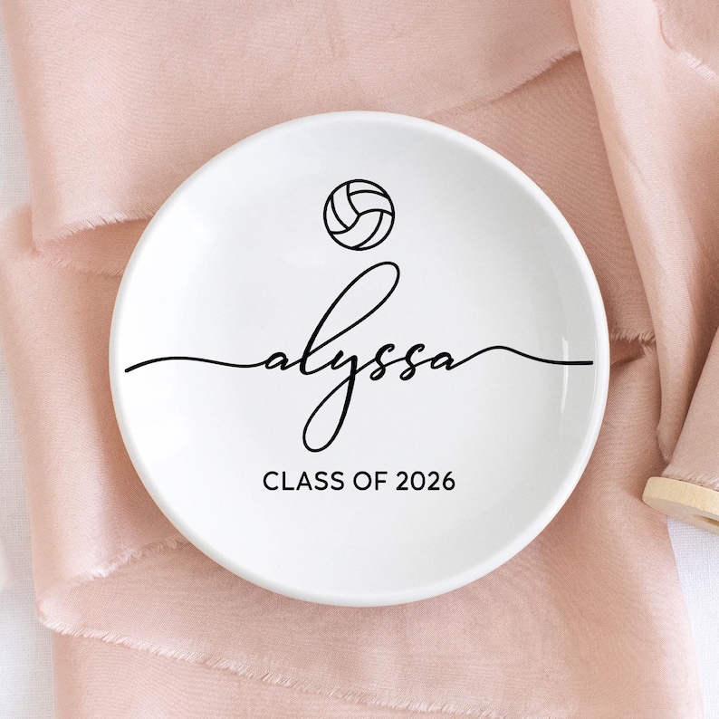 May include: A white ceramic ring dish with a black volleyball graphic and the name "alyssa" in cursive script. Below the name is the text "CLASS OF 2026". The dish is round and sits on a pink fabric surface.