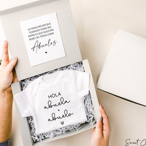 Op de afbeelding: Een witte baby romper met de tekst "HOLA abuela + abuelo" in zwarte schrift. De romper zit in een witte cadeaudoos met een witte kaart waarop staat: "LO &Uacute;NICO MEJOR QUE TENERLOS COMO MIS PAPAS ES QUE NUESTRO BEBE LOS TENGA COMO Abuelos".