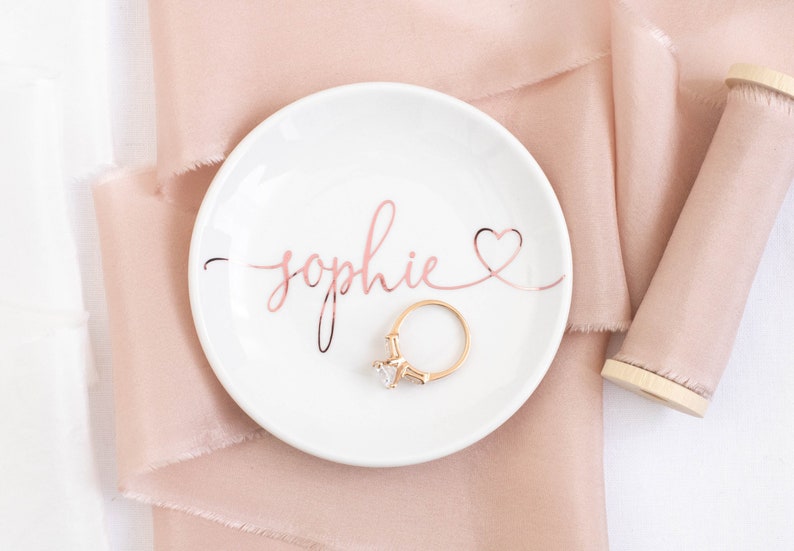 May include: A white ceramic ring dish with the name "Sophie" written in rose gold lettering with a heart at the end. A gold ring with a clear stone is sitting on the dish.