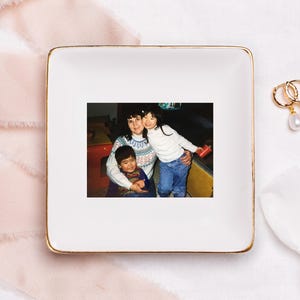 以下が含まれることがあります： 金色の縁が付いた白い正方形のセラミック製の小物入れ。この皿には、女性と2人の子供の写真が額縁に入っています。写真は、家族の瞬間を捉えたヴィンテージのスナップショットです。