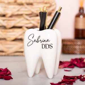 May include: White ceramic tooth-shaped pencil holder with the name "Sabrina DDS" written in black lettering.