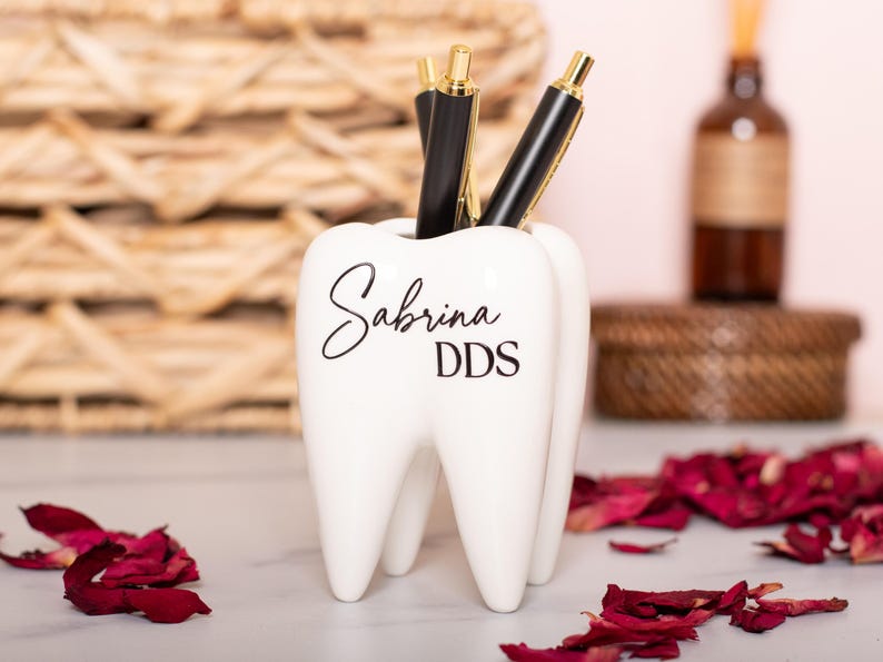 May include: White ceramic tooth-shaped pencil holder with the name "Sabrina DDS" written in black lettering.
