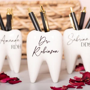 May include: Three white ceramic tooth-shaped pencil holders with black lettering. The holders are personalized with the names "Amanda RDH", "Dr. Robinson", and "Sabrina DDS".