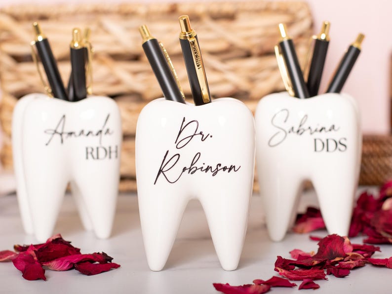 May include: Three white ceramic tooth-shaped pencil holders with black lettering. The holders are personalized with the names "Amanda RDH", "Dr. Robinson", and "Sabrina DDS".