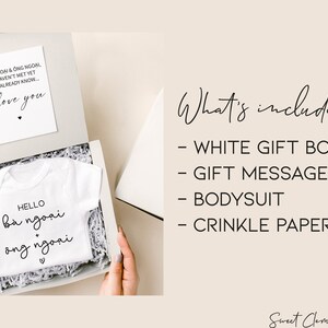 May include: A white gift box containing a white baby bodysuit with Vietnamese text, a card with text, and crinkle paper. The bodysuit says "Hello b&agrave; ngoại + &ocirc;ng ngoại". The card says "I love you".