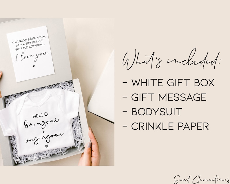 May include: A white gift box containing a white baby bodysuit with Vietnamese text, a card with text, and crinkle paper. The bodysuit says "Hello b&agrave; ngoại + &ocirc;ng ngoại". The card says "I love you".