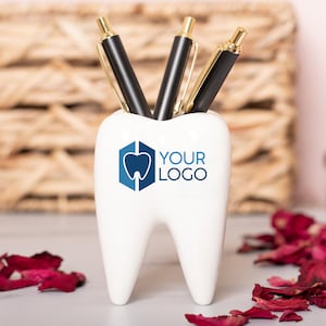 May include: White ceramic tooth-shaped desk organiser with three black pens inside. The organiser has a blue tooth logo and the text "YOUR LOGO" printed on the front.