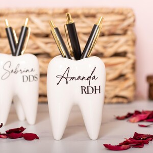 May include: Two white ceramic tooth-shaped pencil holders. The holder on the left has the name "Sabrina DDS" written on it. The holder on the right has the name "Amanda RDH" written on it.