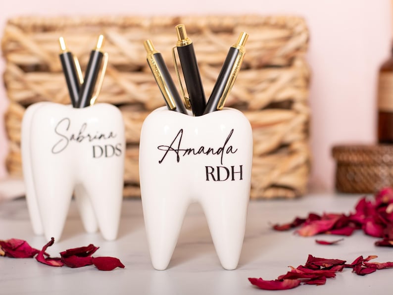 May include: Two white ceramic tooth-shaped pencil holders. The holder on the left has the name "Sabrina DDS" written on it. The holder on the right has the name "Amanda RDH" written on it.