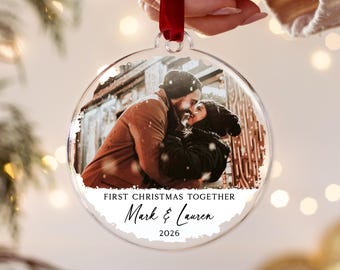 May include: A round, clear acrylic Christmas ornament featuring a printed photograph of a couple embracing in winter attire. The text reads FIRST CHRISTMAS TOGETHER, Mark & Lauren, and 2026. A red ribbon is attached to the top for hanging.