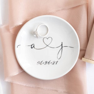 May include: White ceramic ring dish with a silver heart and the initials "a & j" and the date "06-26-21" in cursive script.