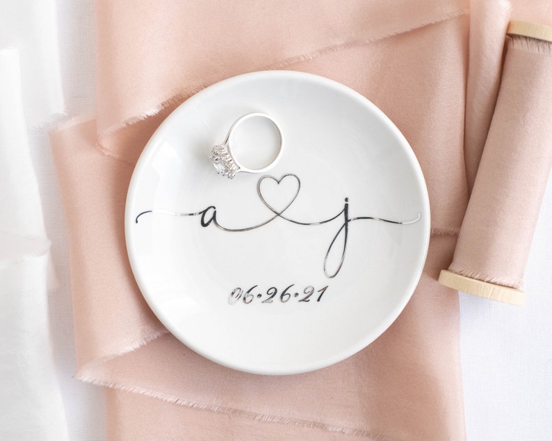 May include: White ceramic ring dish with a silver heart and the initials "a & j" and the date "06-26-21" in cursive script.