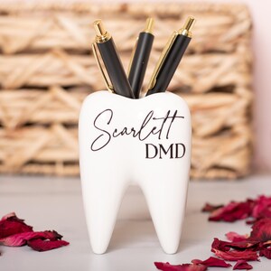 May include: White ceramic tooth-shaped desk organizer with the name "Scarlett DMD" written in black script. Three black pens are in the organizer.