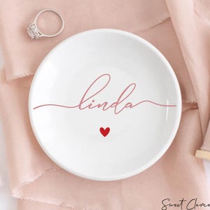 K&ouml;nnte beinhalten: Eine wei&szlig;e Keramik-Ring Schale mit dem Namen "Linda" in rosa Schrift. Ein kleines rotes Herz befindet sich mittig unter dem Namen.