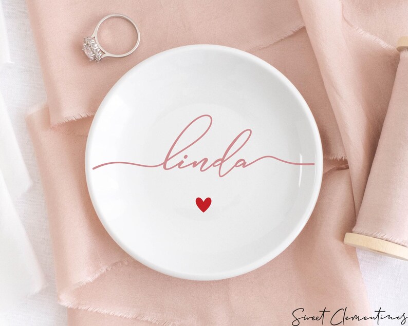 K&ouml;nnte beinhalten: Eine wei&szlig;e Keramik-Ring Schale mit dem Namen "Linda" in rosa Schrift. Ein kleines rotes Herz befindet sich mittig unter dem Namen.
