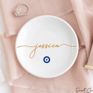 May include: White ceramic ring dish with the name "jessica" written in gold script. A blue and white evil eye charm is centered below the name.