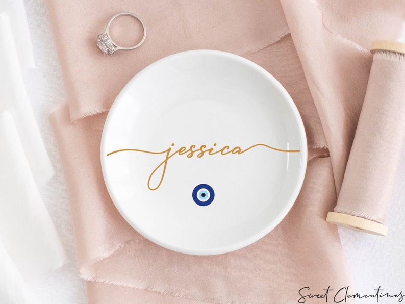 May include: White ceramic ring dish with the name "jessica" written in gold script. A blue and white evil eye charm is centered below the name.