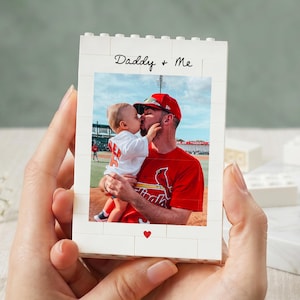 May include: A white brick-style photo frame with the words "Daddy + Me" in black script. The frame holds a color photo of a man kissing a baby. A red heart is printed at the bottom of the frame.