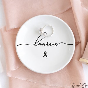 Op de afbeelding: Een witte keramische ringenschotel met zwarte, cursieve tekst die "Lauren" zegt en een zwart lint. Een zilveren ring ligt op de schotel.