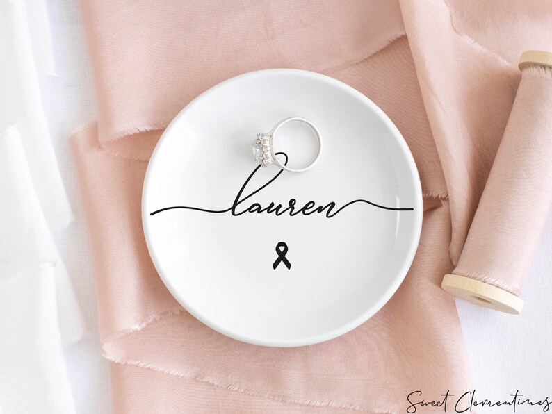 Op de afbeelding: Een witte keramische ringenschotel met zwarte, cursieve tekst die "Lauren" zegt en een zwart lint. Een zilveren ring ligt op de schotel.