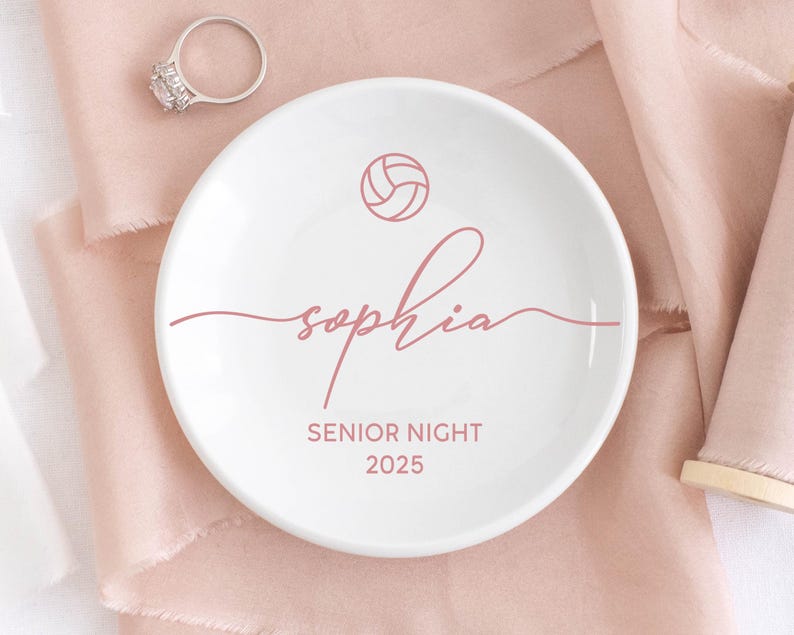May include: White ceramic ring dish with a pink volleyball graphic, the name "Sophia" in cursive, and "Senior Night 2025" printed in pink. A silver ring with a large stone sits nearby.