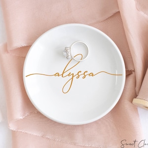 May include: White ceramic ring dish with a gold cursive inscription that reads "alyssa". A silver ring with a clear stone is sitting on the dish.