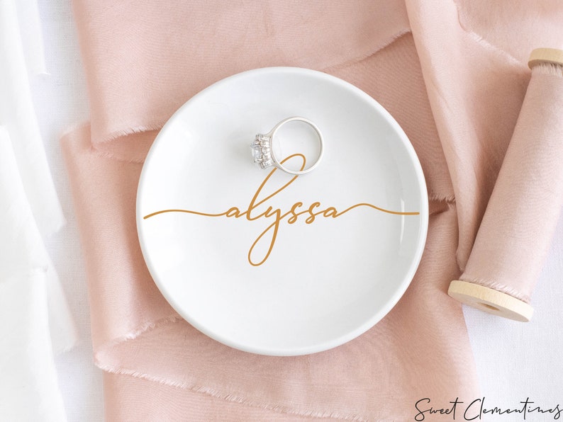 May include: White ceramic ring dish with a gold cursive inscription that reads "alyssa". A silver ring with a clear stone is sitting on the dish.