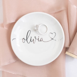 May include: A white ceramic ring dish with the name "olivia" written in silver script and a silver heart. A silver ring is sitting on the dish. The dish is on a pink satin ribbon.