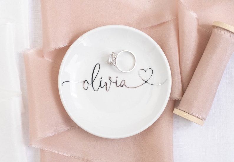 May include: A white ceramic ring dish with the name "olivia" written in silver script and a silver heart. A silver ring is sitting on the dish. The dish is on a pink satin ribbon.
