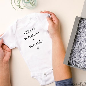 Op de afbeelding: Een witte baby romper met zwarte tekst die "HELLO nana + nani" luidt met een klein hartje.