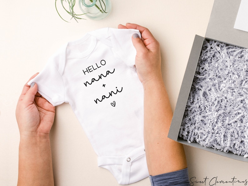Op de afbeelding: Een witte baby romper met zwarte tekst die "HELLO nana + nani" luidt met een klein hartje.