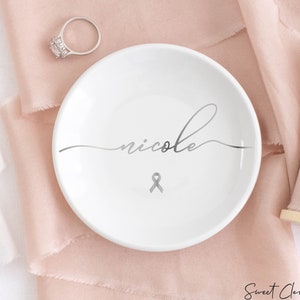 Op de afbeelding: Witte keramische ringenschotel met de naam "nicole" in cursief schrift en een zilveren lint.