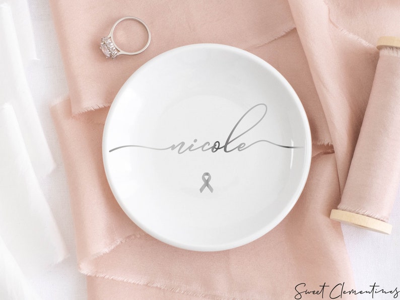 Op de afbeelding: Witte keramische ringenschotel met de naam "nicole" in cursief schrift en een zilveren lint.