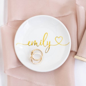 May include: White ceramic ring dish with the name "emily" written in gold script with a gold heart. Two gold rings are sitting in the dish.