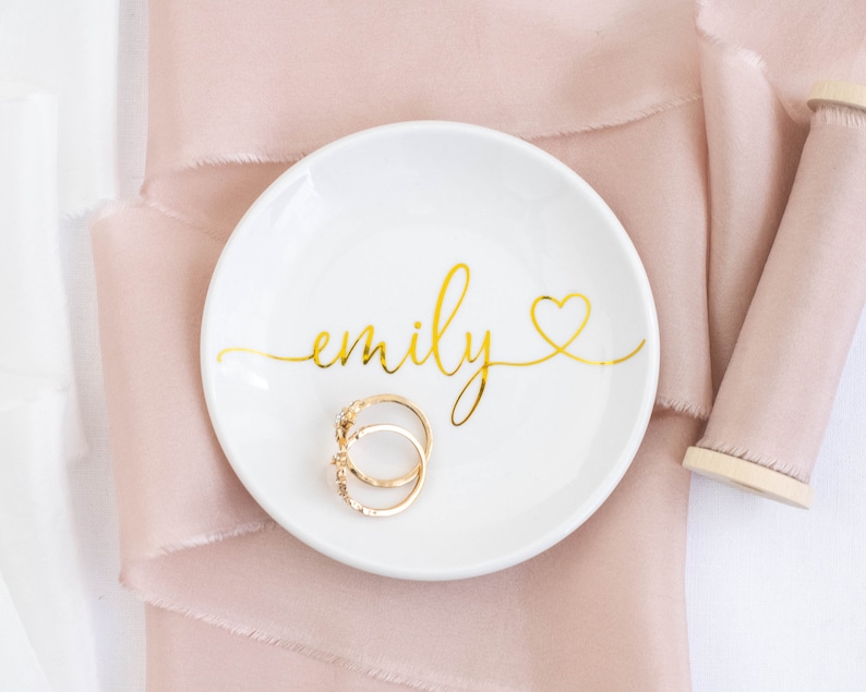 May include: White ceramic ring dish with the name "emily" written in gold script with a gold heart. Two gold rings are sitting in the dish.