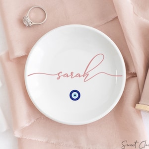 May include: White ceramic ring dish with the name "sarah" written in pink script and a blue evil eye charm in the center.