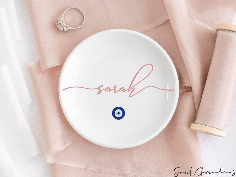 May include: White ceramic ring dish with the name "sarah" written in pink script and a blue evil eye charm in the center.