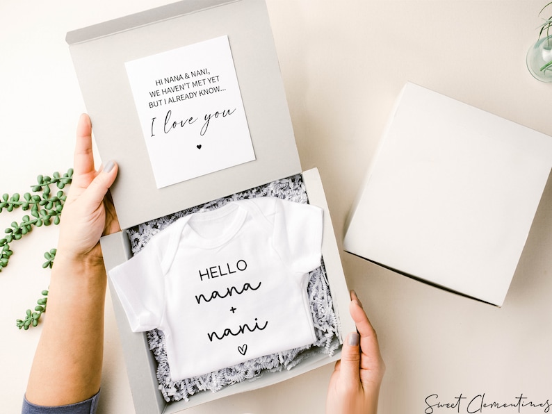 Op de afbeelding: Een witte baby romper met de tekst "HELLO nana + nani" en een klein hartje. De romper zit in een witte cadeaudoos met wit tissuepapier. Een kaart met de tekst "HI NANA & NANI, WE HAVEN'T MET YET BUT I ALREADY KNOW... I love you" ligt op het tissuepapier.