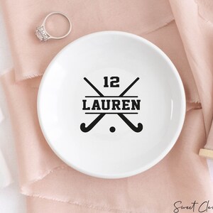 May include: White ceramic ring dish with a black hockey stick design and the text "12 LAUREN".