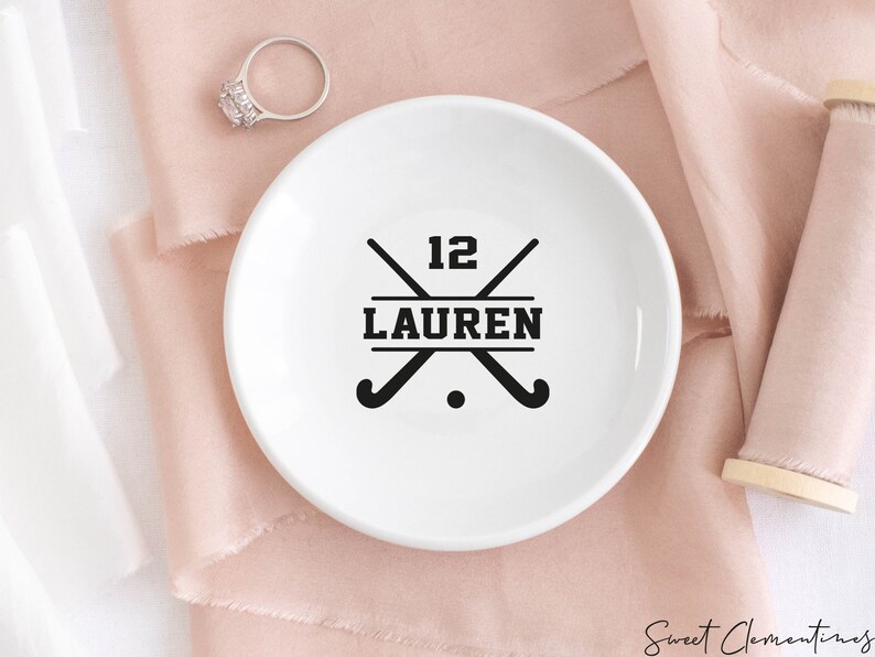 May include: White ceramic ring dish with a black hockey stick design and the text "12 LAUREN".
