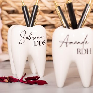 May include: Two white ceramic tooth-shaped pencil holders. The holder on the left has the name Sabrina DDS written on it. The holder on the right has the name Amanda RDH written on it.